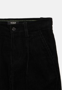 Zwart corduroy broek met knoopsluiting, zijzakken, riemlussen en een zichtbaar "Scotch & Soda" label aan de binnenkant van de taille.