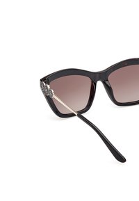 Gafas de sol negras en forma de ojo de gato con lentes marrones degradados, que presentan un elegante herraje floral plateado en las patillas y un acabado brillante y suave.