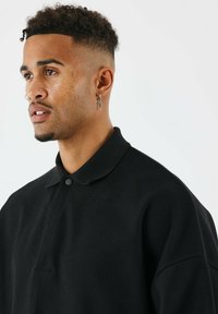 Polo shirt corto a maniche corte di colore nero e texture, con colletto e chiusura a bottone, indossato da una persona con capelli corti e ricci e piercing sul viso.