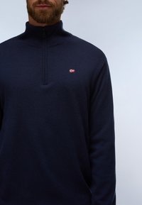 Homme barbu portant un pull bleu marine à col zippé quart et petit logo drapeau rouge, blanc et bleu sur la poitrine, sur fond neutre.