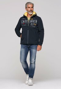 Marinefarbener Reißverschlussjacke mit gelbem Kapuzenfutter und grafischem Text; kombiniert mit hellblauen Jeans und weißen Turnschuhen. Verfügt über vordere Reißverschlusstaschen.