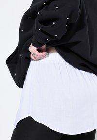 Sudadera negra con acentos de perlas en las mangas, superpuesta sobre una blusa blanca de diseño fluido con dobladillo asimétrico. Leggings negros visibles en la parte inferior.