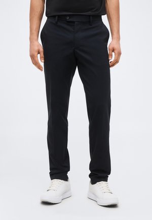 Man wearing black slim-fit trousers and white sneakers, standing against a plain light background.