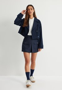 Giacca doppiopetto cropped blu navy con dettagli in bottone, abbinata a una gonna plissettata coordinata con tasche frontali e calze sopra il ginocchio blu navy.