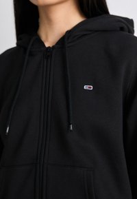 Haut à zip noir en tissu doux, avec une poche avant, une capuche à cordon de serrage et un petit logo coloré sur la poitrine gauche.