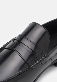 Mocasín de piel negra que presenta un diseño sin costuras, costuras sutiles y un acabado suave y brillante, con un detalle de tira ancha en la parte frontal.