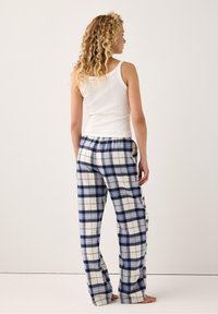 Kvinna med lockigt hår, iklädd en vit linne och blå, vit och svart rutiga pyjamasbyxor, stående barfota mot en enkel bakgrund.