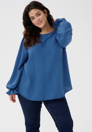 Blaues Langarmshirt aus strukturiertem Material, lockere Passform und geraffte Ärmelbündchen. Kombiniert mit dunklen Jeans und rundem Ausschnitt.