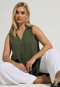 Top verde senza maniche, con colletto e tessuto texturizzato, chiusura con bottoni e vestibilità rilassata, abbinato a pantaloni bianchi ampi.