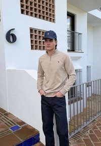 Mladý muž ve světle hnědém svetru s krátkým zipem, tmavě modré čepici a tmavých džínách stojí před bílou budovou u čísla 6 a kovového zábradlí.