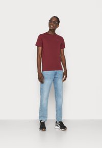 Bordeaux t-shirt met korte mouwen en een ronde hals, gecombineerd met lichtblauwe jeans en zwarte sneakers, staand tegen een neutrale achtergrond.