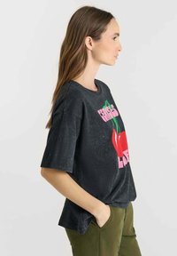 Mujer con cabello largo castaño que lleva una camiseta negra de gran tamaño con un gráfico de cerezas rojas y texto rosa, combinada con pantalones verde oliva, con las manos en los bolsillos.