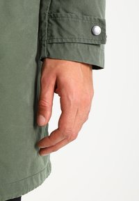 Manche de veste extérieure verte avec fermeture à bouton, fabriquée dans un tissu lisse. Une main repose le long de la manche, montrant le ton de peau et la texture.
