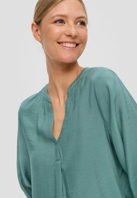 Licht teal blouse met een V-hals, geplooide details en een ontspannen pasvorm. Gemaakt van zachte, gestructureerde stof met lange, luchtige mouwen.