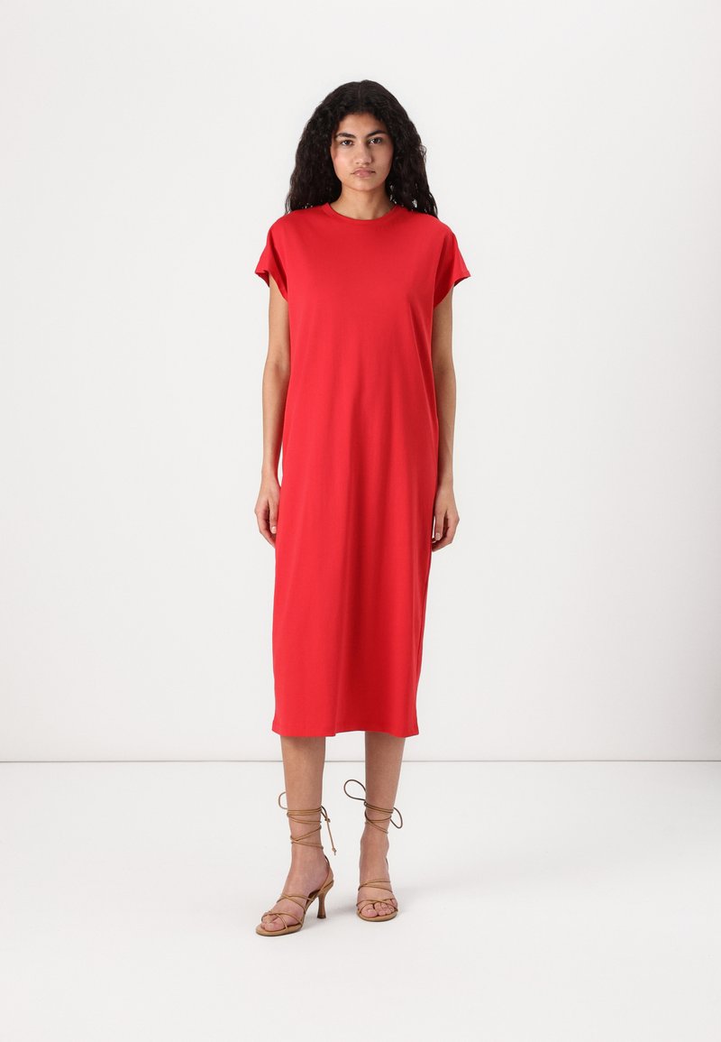 Vestido vermelho, até ao joelho, em tecido liso, com mangas curtas e um corte descontraído. Usam-se sandálias altas, de salto, em tom bege, com tiras.