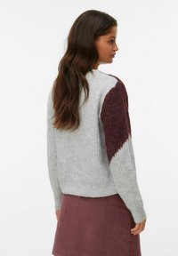 Szary sweter wykonany na drutach z bordową teksturowaną łatą na lewym ramieniu. Posiada okrągły dekolt i ściągacz na dole, zestawiony z bordową spódnicą.