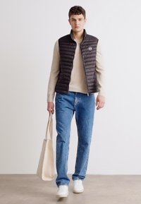 Gilet trapuntato nero, maglione beige, jeans azzurri chiari e sneaker bianche. Borsa tote beige in mano. Design semplice, texture casual.
