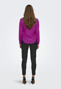 Blusa de satén fucsia de manga larga, combinada con pantalones negros ajustados y zapatos de tacón alto, vista desde la parte posterior.