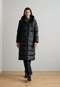 Langer schwarzer Daunenmantel mit Kapuze, mit gesteppten Abschnitten, Reißverschluss und zwei Seitentaschen. Das Modell trägt schwarze Hosen und Stiefeletten.