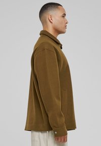 Brown fleece jacket with a collar, side pockets, and buttoned cuffs. The design is relaxed-fit, featuring a smooth texture and solid color.