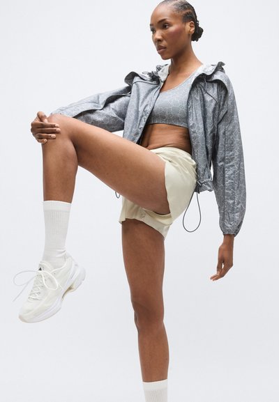 Ανοιχτό γκρι μπουφάν με κουκούλα με βολάν, αθλητικό μπουστάκι με σχέδια και μπεζ σορτς. Λευκά αθλητικά παπούτσια με χοντρές σόλες και λευκές κάλτσες μέχρι τον αστράγαλο.