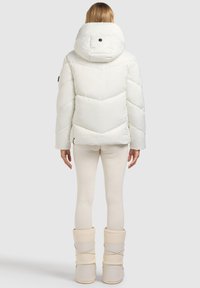 Biała kurtka puchowa z wysokim kołnierzem, przeszyciami w kształcie szewrona i czarnym ozdobnym guzikiem. Do tego jasnobeżowe legginsy i buty.