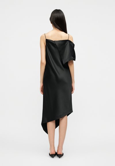 Γυναίκα με μακριά σκούρα μαλλιά, που φοράει ασύμμετρο μαύρο σατέν φόρεμα με ένα μανίκι εκτός ώμου και μαύρα παπούτσια με τακούνι, στέκεται γυρισμένη με την πλάτη.