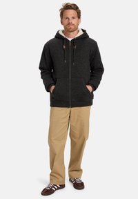 Uomo in piedi con le mani nelle tasche, indossa un felpa con zip scura foderata in sherpa, pantaloni beigi e scarpe da ginnastica marroni con strisce bianche.