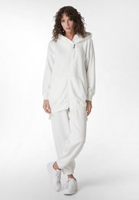Sudadera blanca con cremallera y capucha grande, bolsillos frontales y puños de canalé, combinada con pantalones jogger blancos a juego y zapatillas blancas.