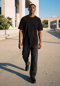 Tânăr îmbrăcat într-un tricou negru și pantaloni merge pe afară într-o zi însorită, ținând o sticlă cu apă în mâna dreaptă.