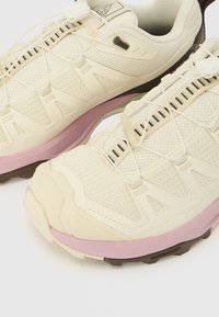 Αθλητικά παπούτσια σε ανοιχτό μπεζ χρώμα με ανώτερο μέρος από διχτυωτό υλικό, ενισχυμένες λεπτομέρειες και ροζ ενδιάμεση σόλα· διαθέτουν κομψή μορφή και ελαστικό σύστημα κορδονιών.