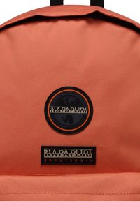 Zaino in tessuto arancione con un patch nero rotondo con un design del globo e un'etichetta nera rettangolare con il testo "NAPAPIJRI geographic".