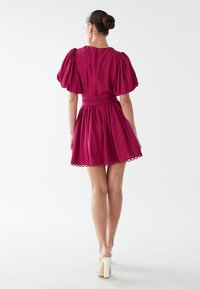 Růžové šaty s volnými rukávy, uplým pasem, rozšířenou sukní a vroubkovaným lemem, vyrobené z hladké látky.