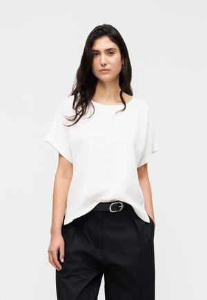 Valkoinen lyhythihainen paita, joka on valmistettu sileästä kankaasta ja jossa on rento istuvuus. Yhdistetty korkeavyötäröisiin mustiin housuihin ja mustaan vyöhön, jossa on hopeanvärinen solki.