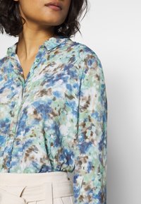 Floral blouse with a blend of blue, green, and brown hues. It features a collared neckline and button closure, made from lightweight fabric.