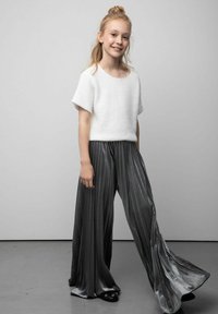 Biały, teksturowany top z krótkim rękawem w połączeniu z błyszczącymi, plisowanymi, szerokimi spodniami w ciemnoszarym kolorze. Czarne wsuwane buty z ozdobą w kształcie oka.