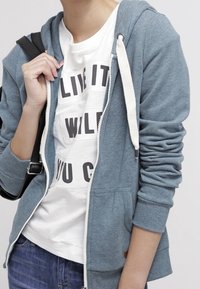 Felpa zip blu chiaro con coulisse bianche, tasche frontali e polsini a coste, indossata sopra una t-shirt bianca con grafica e jeans.