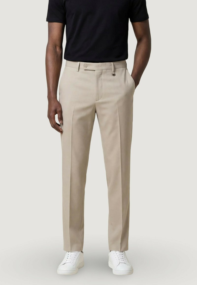 Man wearing beige tailored trousers, black short-sleeve shirt, and white sneakers, standing with one hand in pocket.