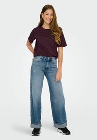 Jeans blu larghi con orli arrotolati, abbinati a una maglietta a maniche corte di colore viola scuro. L'outfit è completato da sneaker nere.