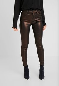 Metaliczne, brązowe obcisłe jeansy o gładkiej fakturze i wysokim stanie, w połączeniu z czarną koszulą z długim rękawem i czarnymi kozakami za kolano.