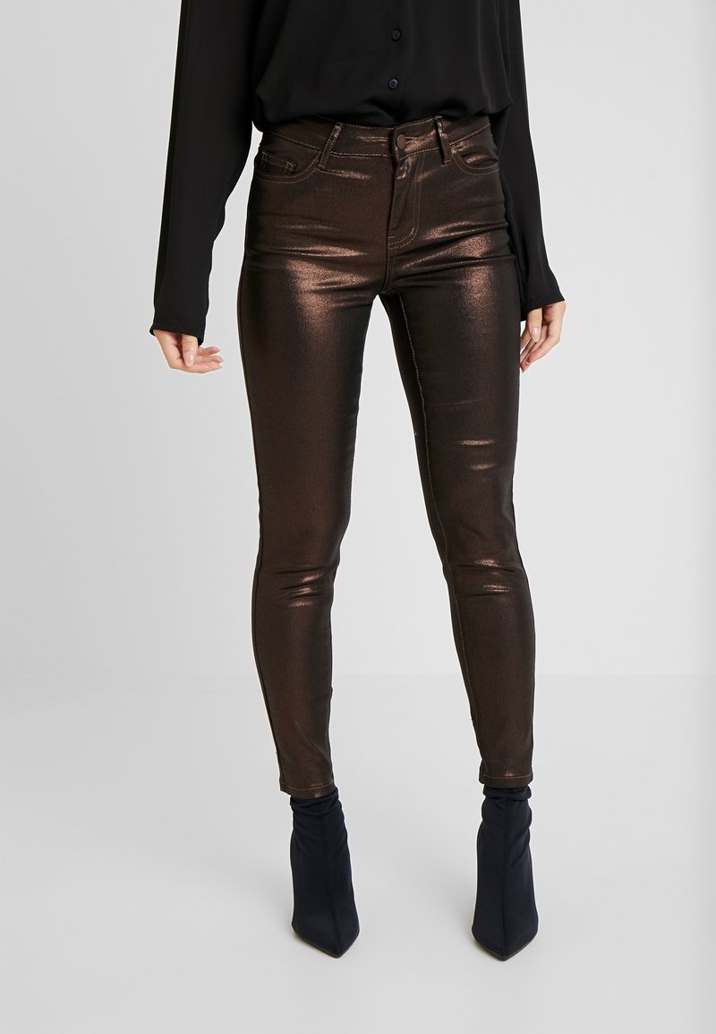 Metaliczne, brązowe obcisłe jeansy o gładkiej fakturze i wysokim stanie, w połączeniu z czarną koszulą z długim rękawem i czarnymi kozakami za kolano.