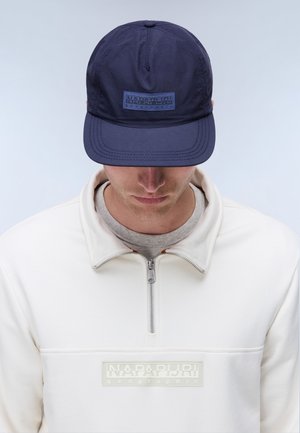 Homme portant une casquette bleu marine et un sweat-shirt blanc zippé, tous deux avec le logo de la marque Napapijri Geographic.