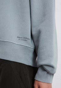 Lichtblauwe sweatshirt van zacht materiaal, met ribgebreide boorden en onderrand. Logo in donkere tekst in de linksonderhoek.