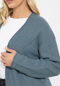 Gilet en maille bleu ardoise, présentant un design en v, une texture côtelée et de petits trous décoratifs sur les manches.