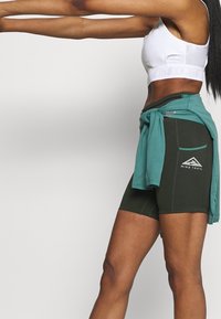 Vrouw in witte sportbeha, zwarte Nike Trail-short, met een teal jasje om haar taille gebonden, steekt haar armen naar voren.