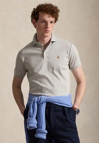 Jeune homme portant un polo gris clair avec un logo, un pantalon bleu marine et un pull bleu noué autour de la taille, debout les mains dans les poches.