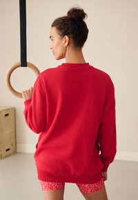 Röd sweatshirt med ribbade muddar och fåll, med en logotyp på ryggen. Modellen håller i en trägymringar och bär mönstrade shorts under.