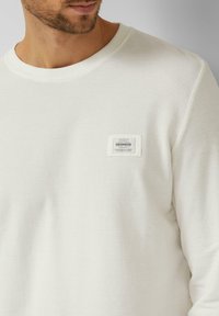 Witte sweatstof met een structuur, voorzien van een ronde hals en een klein patchlogo op de linkerborst. De stof lijkt zacht en gebreid.
