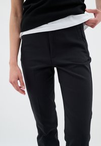 Černé kalhoty s přesným střihem a hladkou texturou, slim fit, a jemnými předními záhyby, v kombinaci s bílým vrstveným topem.