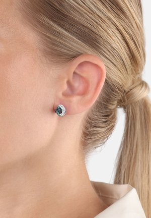 Silberne Ohrstecker mit einem kreisförmigen Design, ausgestattet mit schwarzen und türkisfarbenen Steinen sowie einer strukturierten Oberfläche, die am Ohr eines Models getragen werden.